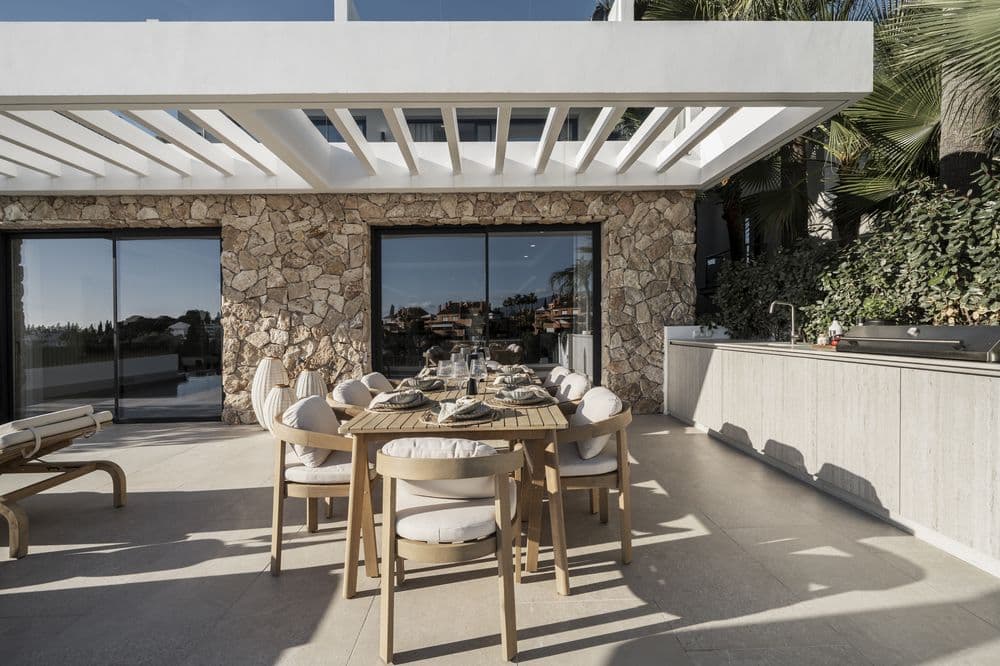Outdoor dining area under a pergola against a stone wall, table set for guests